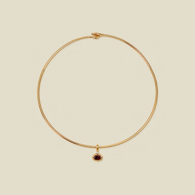 A delicate gold wire circle necklace with a small oval dark red gemstone pendant, displayed on a solid light beige background.