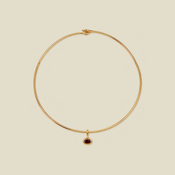 A delicate gold wire circle necklace with a small oval dark red gemstone pendant, displayed on a solid light beige background.