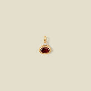 A gold oval pendant charm with a red bezel-set oval gemstone and a clear accent stone on top, set against a solid beige background.