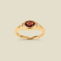 A gold signet ring with a red center oval bezel-set gemstone, and two small clear accent stones on either side, set against a solid beige background.