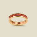A gold eternity ring featuring a continuous channel setting of baguette-cut orange-red gemstones, shown on a solid light beige background.