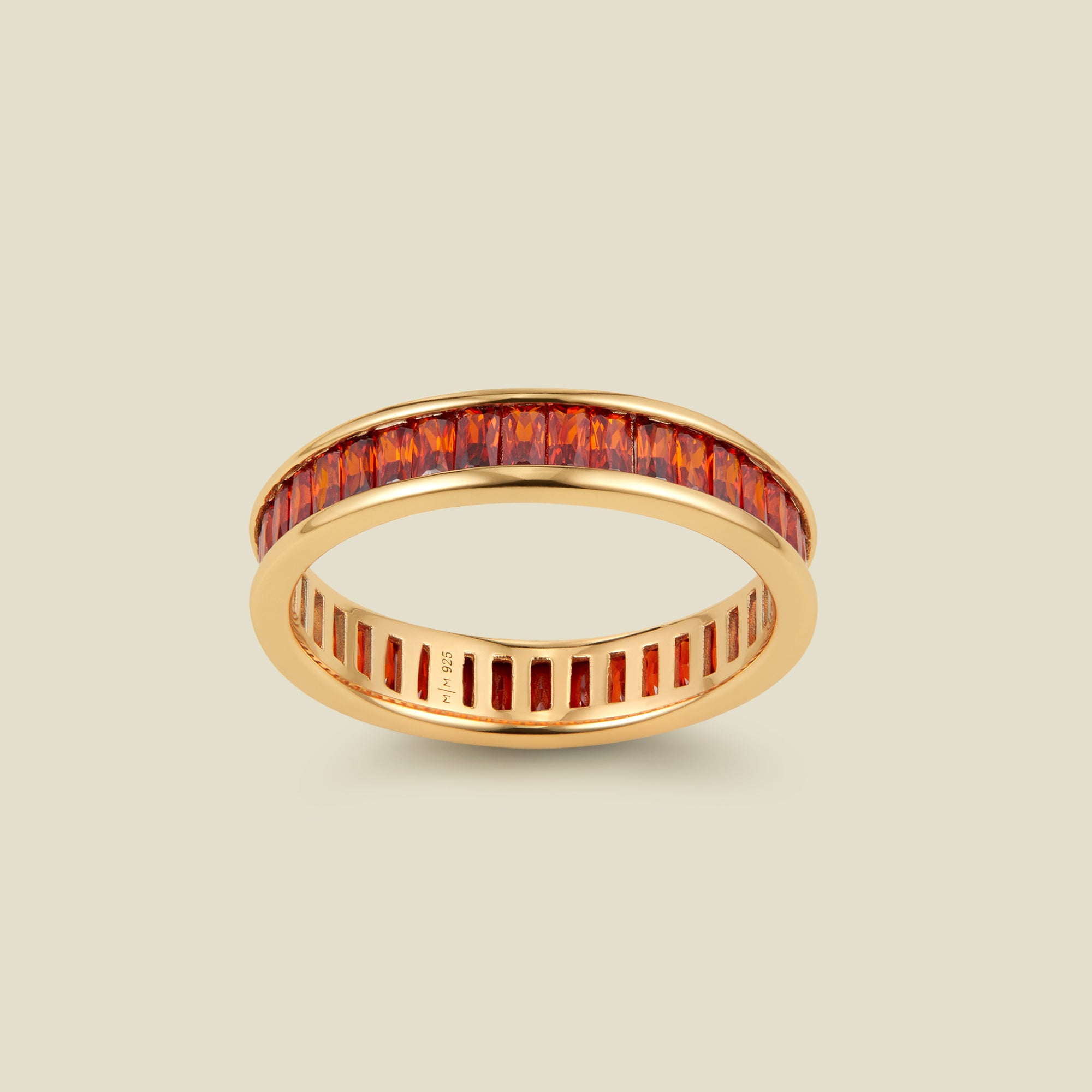 A gold eternity ring featuring a continuous channel setting of baguette-cut orange-red gemstones, shown on a solid light beige background.