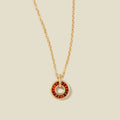 A gold chain necklace with a circular pendant featuring red baguette-cut stones around a hollow center, displayed against a solid light beige background.
