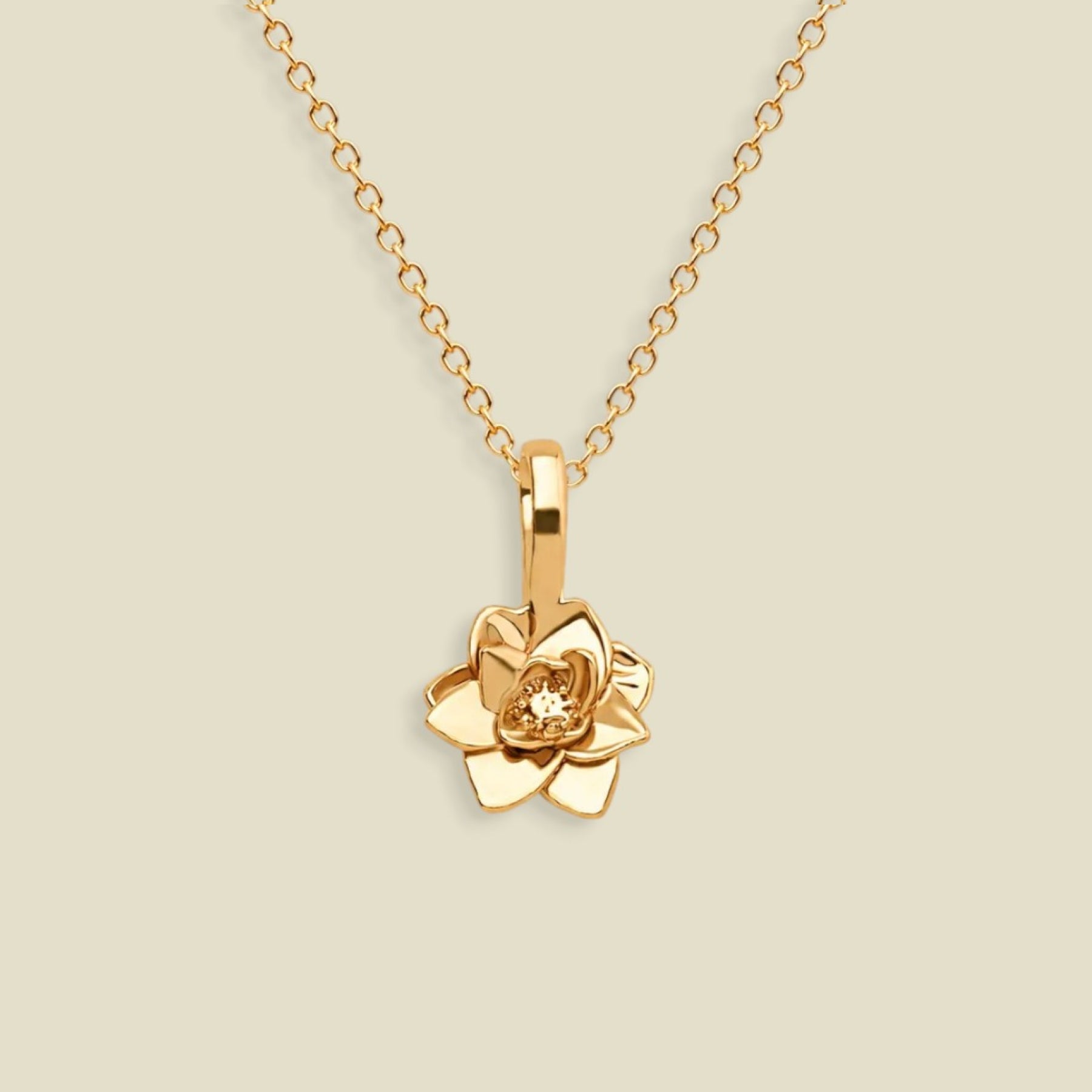 A gold pendant necklace featuring a stylized lotus flower charm, suspended from a delicate gold chain against a solid light beige background.