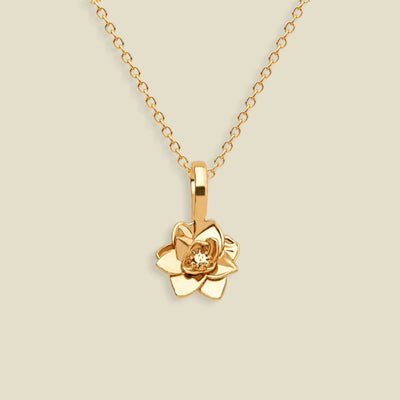 A gold pendant necklace featuring a stylized lotus flower charm, suspended from a delicate gold chain against a solid light beige background.
