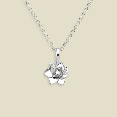 A silver pendant necklace featuring a stylized lotus flower charm, suspended from a delicate silver chain against a solid light beige background.