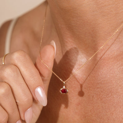 A close-up lifestyle shot of a model wearing a delicate gold chain necklace with a small oval red gemstone pendant, with her hand gently touching the chain.