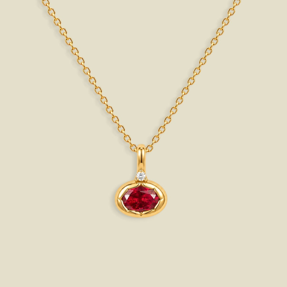 A gold chain necklace with a pendant featuring a bezel-set oval red gemstone and a small clear accent stone above it, shown on a light beige background.