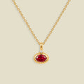 A gold chain necklace with a pendant featuring a bezel-set oval red gemstone and a small clear accent stone above it, shown on a light beige background.