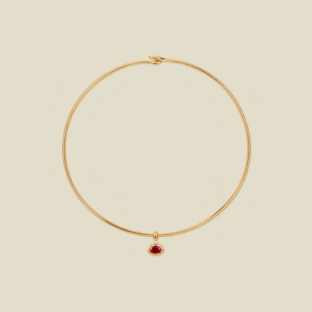 A delicate gold wire choker necklace with a small teardrop-shaped red gemstone pendant, displayed on a solid light beige background.