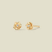A pair of gold lotus flower stud earrings, each featuring a small round clear gemstone at the center, displayed on a solid light beige background.