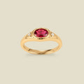 A gold signet ring with a red center oval bezel-set gemstone, and two small clear accent stones on either side, set against a solid beige background.