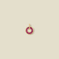 A circular gold pendant featuring red baguette-cut stones around a hollow center, displayed against a solid light beige background.