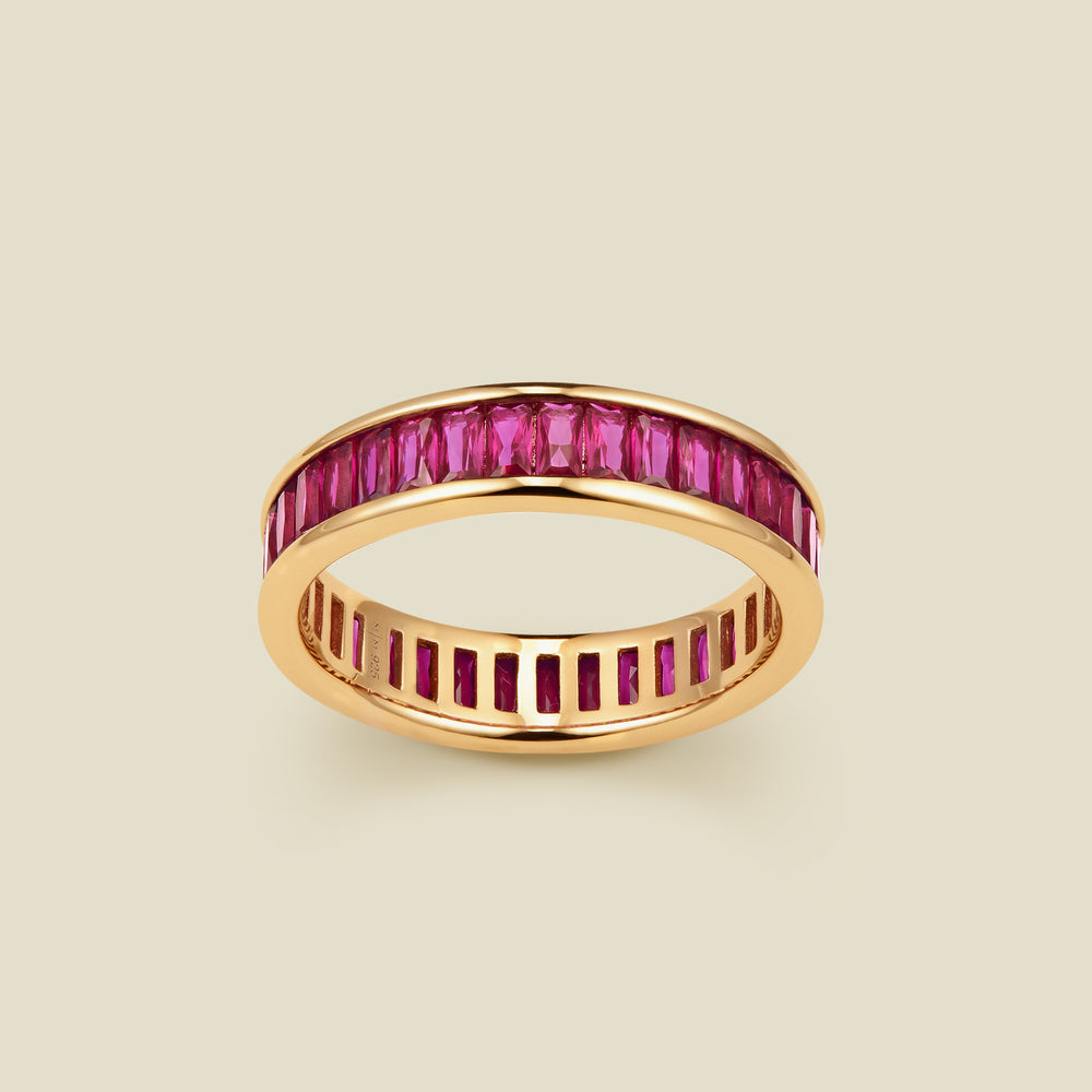 A gold eternity ring featuring a continuous channel setting of baguette-cut red gemstones, shown on a solid light beige background.