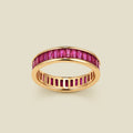 A gold eternity ring featuring a continuous channel setting of baguette-cut red gemstones, shown on a solid light beige background.