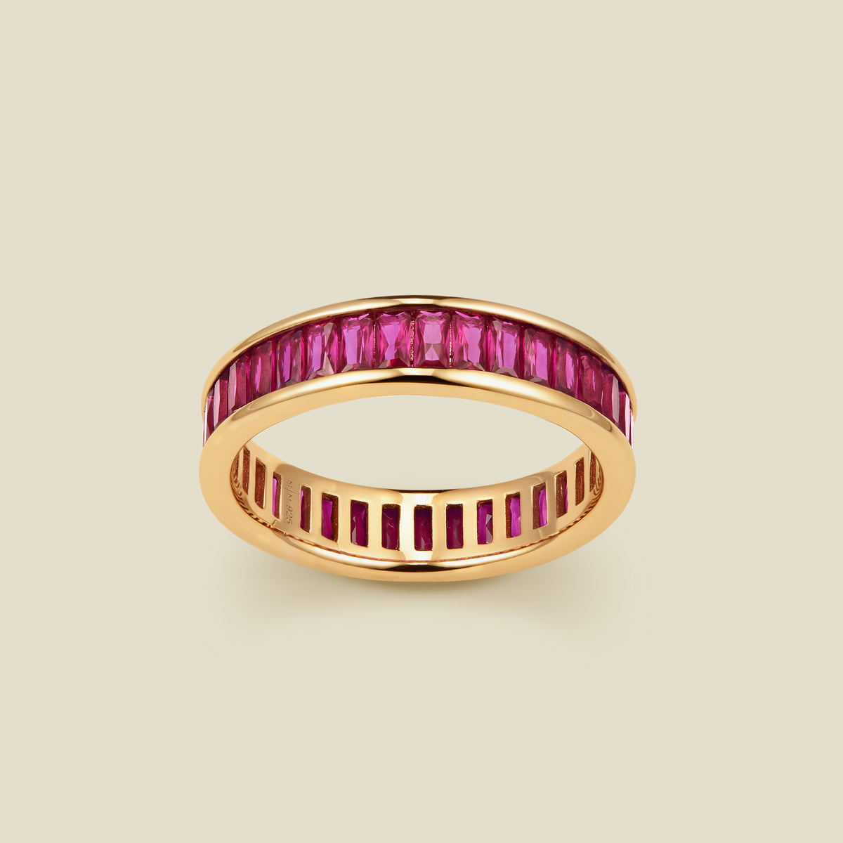 A gold eternity ring featuring a continuous channel setting of baguette-cut red gemstones, shown on a solid light beige background.