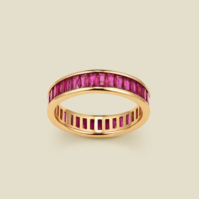 A gold eternity ring featuring a continuous channel setting of baguette-cut red gemstones, shown on a solid light beige background.