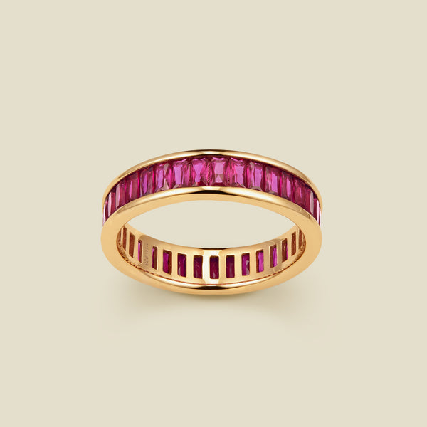 A gold eternity ring featuring a continuous channel setting of baguette-cut red gemstones, shown on a solid light beige background.