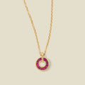 A gold chain necklace with a circular pendant featuring red baguette-cut stones around a hollow center, displayed against a solid light beige background.