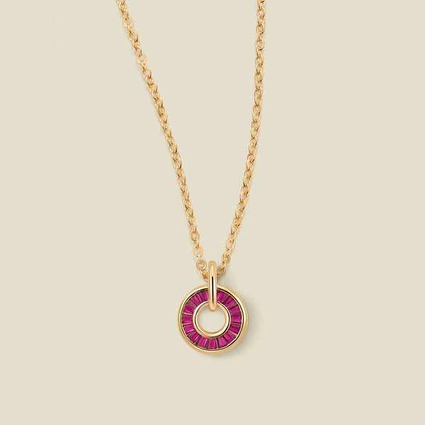 A gold chain necklace with a circular pendant featuring red baguette-cut stones around a hollow center, displayed against a solid light beige background.