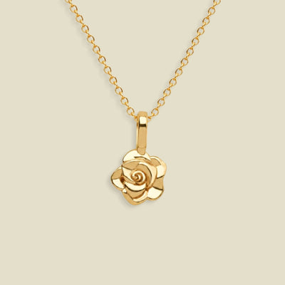 A gold rose pendant necklace on a delicate gold chain, set against a solid light beige background.