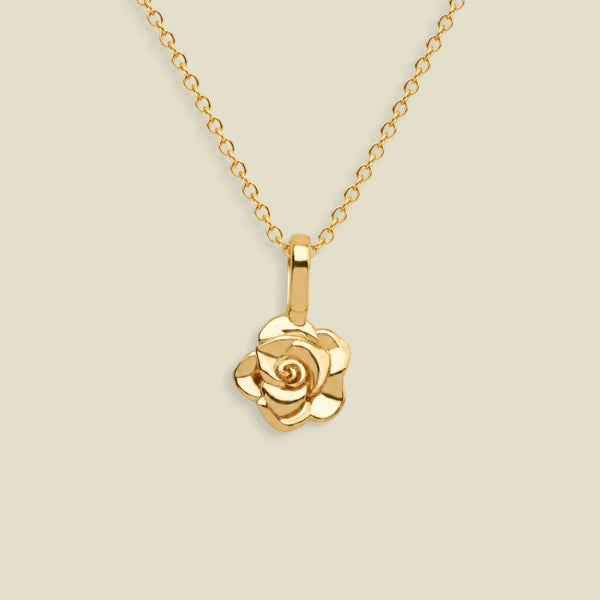A gold rose pendant necklace on a delicate gold chain, set against a solid light beige background.