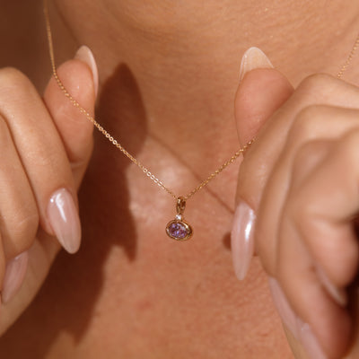 A close-up lifestyle shot of a person wearing a delicate gold chain necklace with a small oval pendant featuring a purple gemstone, held gently by both hands against their chest.