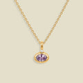 A gold chain necklace with a small oval pendant featuring a bezel set lavender-purple gemstone and a tiny clear accent stone above it, shown on a light beige background.