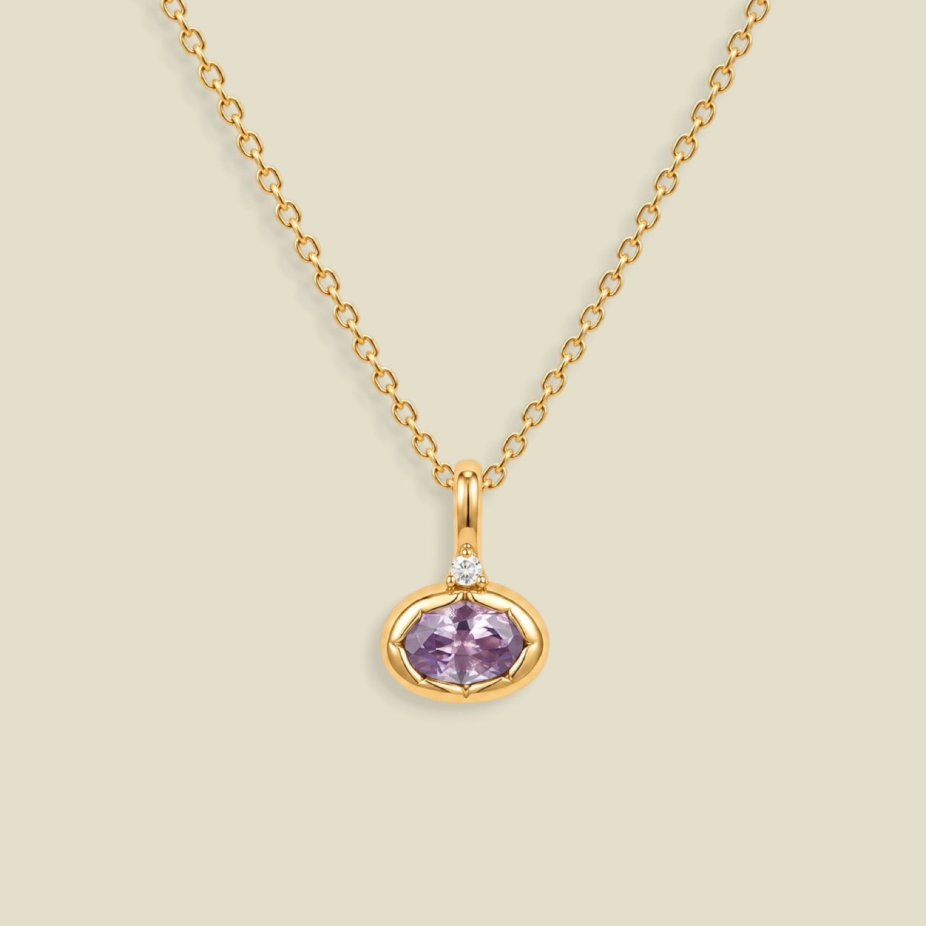 A gold chain necklace with a small oval pendant featuring a bezel set lavender-purple gemstone and a tiny clear accent stone above it, shown on a light beige background.