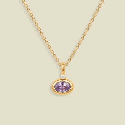 A gold chain necklace with a small oval pendant featuring a bezel set lavender-purple gemstone and a tiny clear accent stone above it, shown on a light beige background.