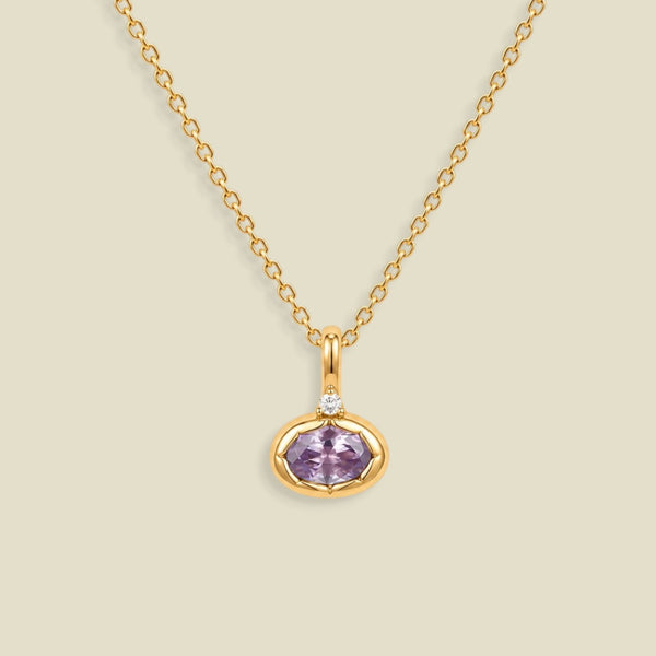 A gold chain necklace with a small oval pendant featuring a bezel set lavender-purple gemstone and a tiny clear accent stone above it, shown on a light beige background.