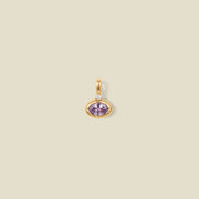 A gold oval pendant charm with a light purple bezel-set oval gemstone and a clear accent stone on top, set against a solid beige background.