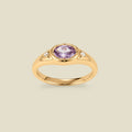 A gold signet ring with a light purple center oval bezel-set gemstone, and two small clear accent stones on either side, set against a solid beige background.