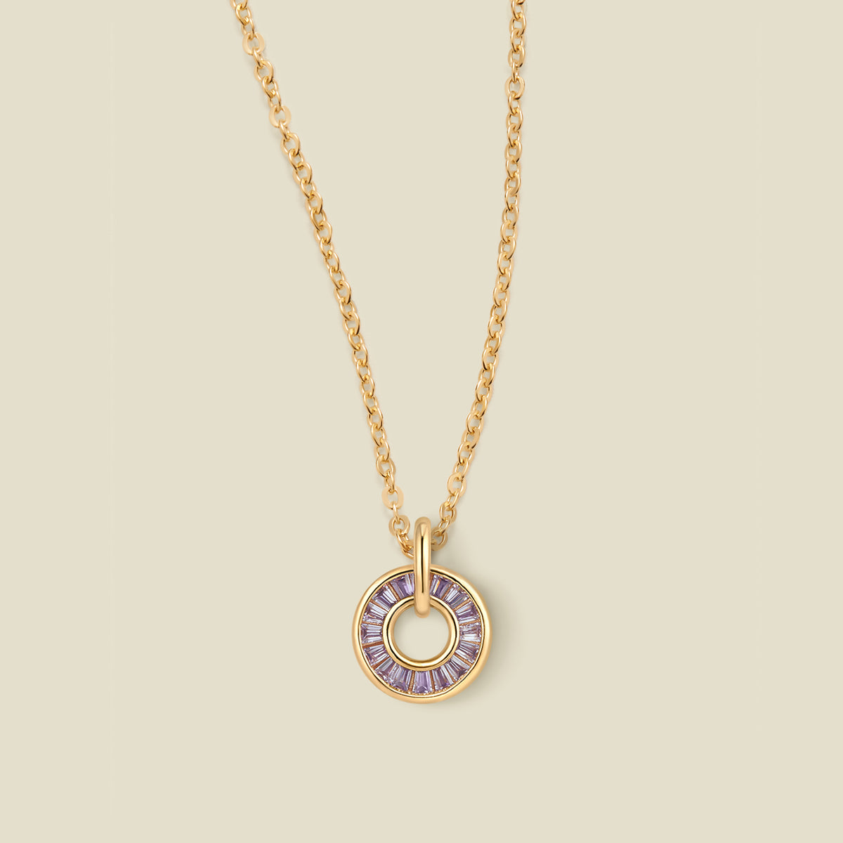 A gold chain necklace with a circular pendant featuring light purple baguette-cut stones around a hollow center, displayed against a solid light beige background.