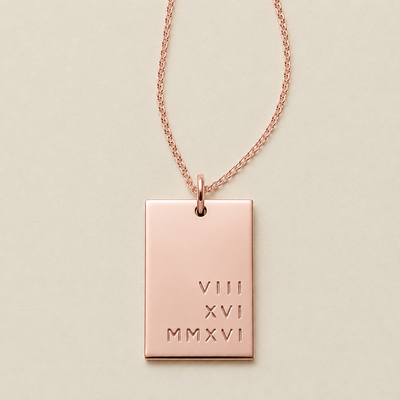 A rose gold rectangular pendant necklace engraved with Roman numerals stacked in three rows on the bottom left quadrant, suspended from a delicate rose gold chain against a light beige background.