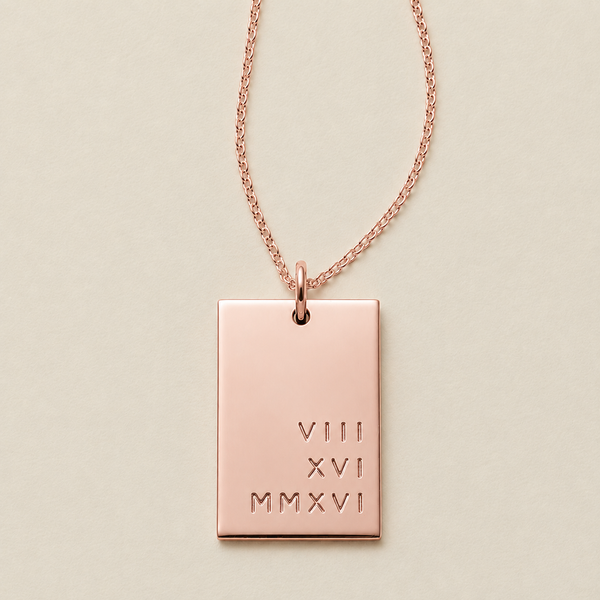 A rose gold rectangular pendant necklace engraved with Roman numerals stacked in three rows on the bottom left quadrant, suspended from a delicate rose gold chain against a light beige background.