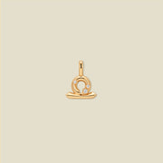 A gold pendant shaped like the Libra zodiac symbol, featuring three small clear gemstones, centered on a solid light beige background.