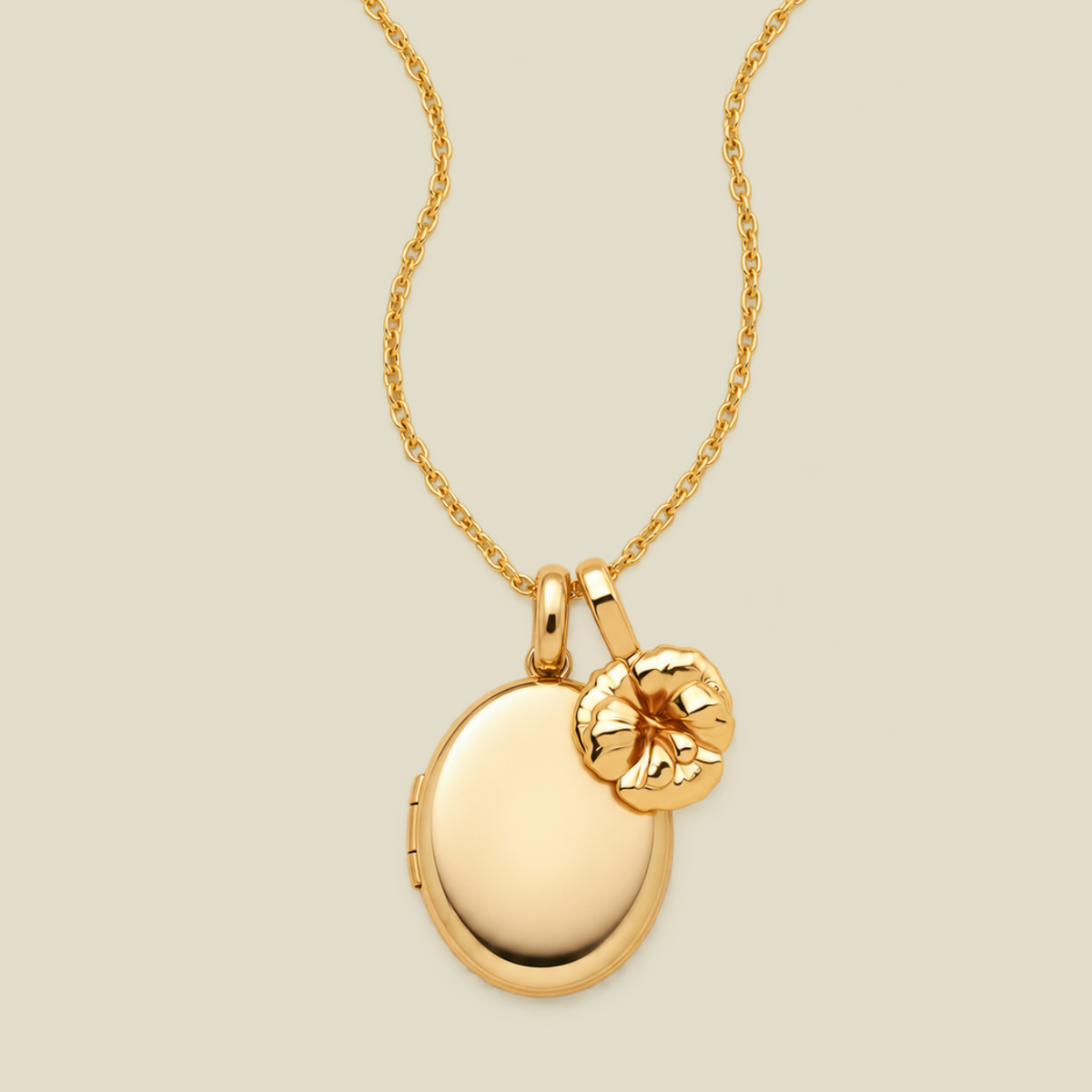 A gold locket necklace with an oval locket pendant and a small textured violet flower charm, shown on a light beige background.