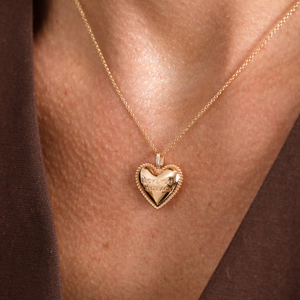 A close-up of a person's neck wearing a delicate gold chain necklace with a heart-shaped pendant with the words "I LOVE YOU, SAY IT BACK". The pendant features a smooth center and a textured, beaded border.