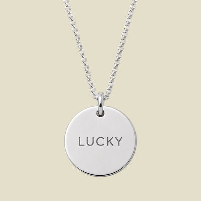 A silver round disc pendant necklace engraved with the word 'LUCKY' in a clean sans-serif font, suspended from a matching silver chain against a solid light beige background.