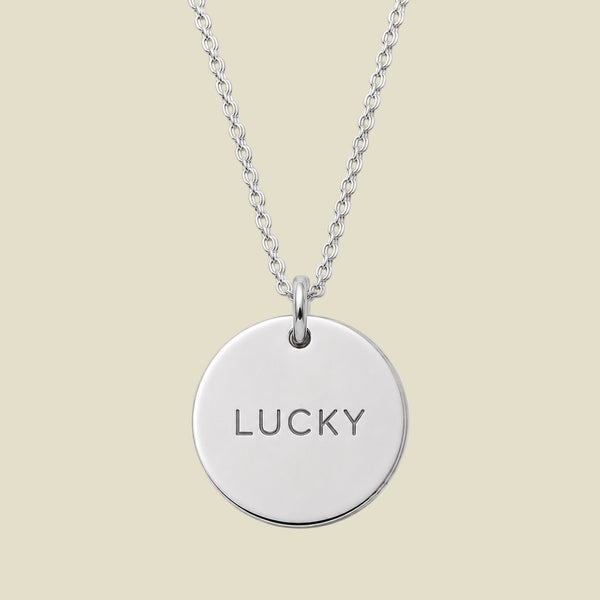 A silver round disc pendant necklace engraved with the word 'LUCKY' in a clean sans-serif font, suspended from a matching silver chain against a solid light beige background.
