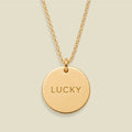 A gold chain necklace with a round gold disc pendant engraved with the word 'LUCKY' in capital letters, shown against a solid light beige background.