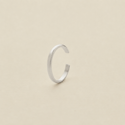 A minimalist silver open-ended ear cuff, shaped like a lowercase letter 'c', standing upright on a plain beige surface.