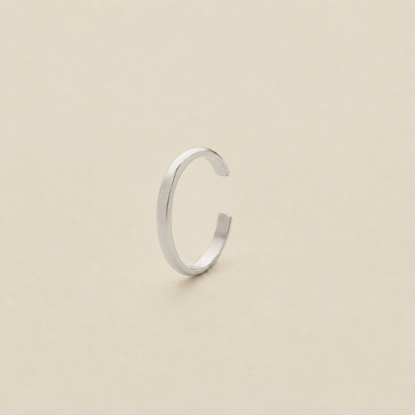 A minimalist silver open-ended ear cuff, shaped like a lowercase letter 'c', standing upright on a plain beige surface.