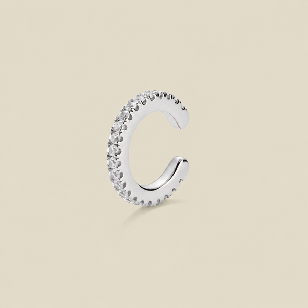 A silver-toned C-shaped huggie hoop ear cuff encrusted with clear cubic zirconia stones, shown on a solid beige background.