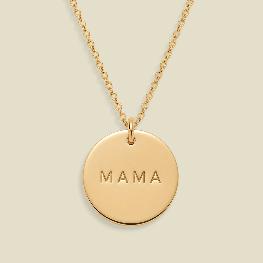 A gold round pendant necklace engraved with the word 'MAMA' in capital letters, suspended from a delicate gold chain against a solid light beige background.