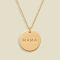A gold round pendant necklace engraved with the word 'MAMA' in capital letters, suspended from a delicate gold chain against a solid light beige background.