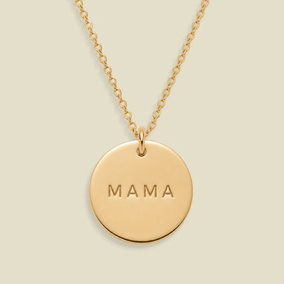 A gold round pendant necklace engraved with the word 'MAMA' in capital letters, suspended from a delicate gold chain against a solid light beige background.