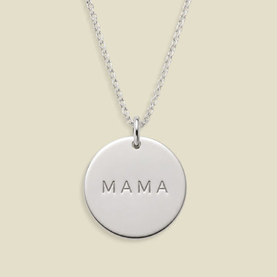 A silver round disc pendant necklace engraved with the word 'MAMA', suspended from a delicate silver chain against a solid light beige background.