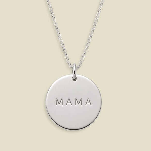A silver round disc pendant necklace engraved with the word 'MAMA', suspended from a delicate silver chain against a solid light beige background.